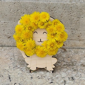 May include: A wooden lion decoration featuring a wreath of vibrant yellow dandelion flowers. The lion's face is cut out of the wood, depicting a smiling expression. The decoration is placed on a concrete surface.