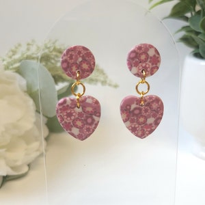 May include: A pair of pink heart-shaped dangle earrings with a floral pattern. The earrings have a small round disc at the top and a gold-colored chain connecting the disc to the heart.