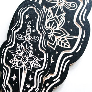 May include: Two black and white woodblock prints of a dagger with a flower design. The prints are in a wavy, cloud-like shape.