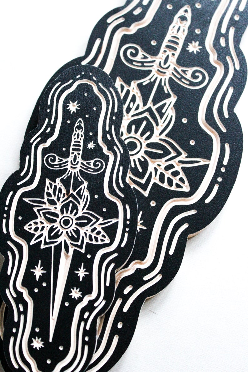 May include: Two black and white woodblock prints of a dagger with a flower design. The prints are in a wavy, cloud-like shape.
