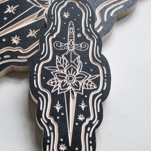 May include: A black and white wood-carved design of a dagger piercing a flower with a star-filled background.