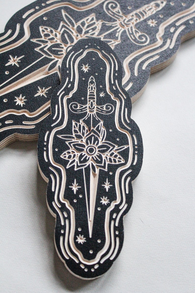 May include: A black and white wood-carved design of a dagger piercing a flower with a star-filled background.