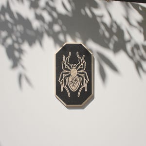 May include: A black octagonal wooden sign with a white spider design. The spider has a detailed body and eight legs.
