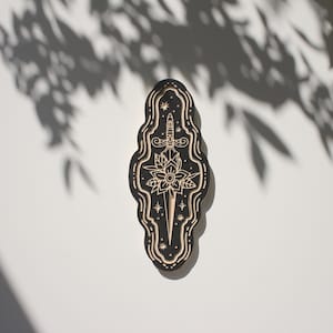 May include: A black wooden plaque with a detailed carving of a dagger piercing a flower. The carving is surrounded by a wavy border and stars.
