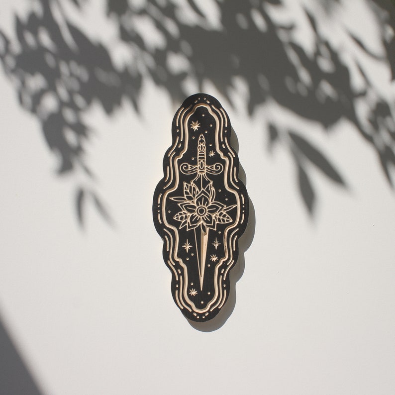 May include: A black wooden plaque with a detailed carving of a dagger piercing a flower. The carving is surrounded by a wavy border and stars.