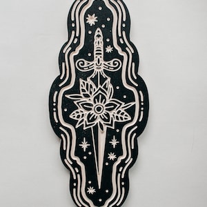 May include: A black and white wooden wall hanging with a stylized dagger and flower design. The dagger is surrounded by a swirling border and stars.