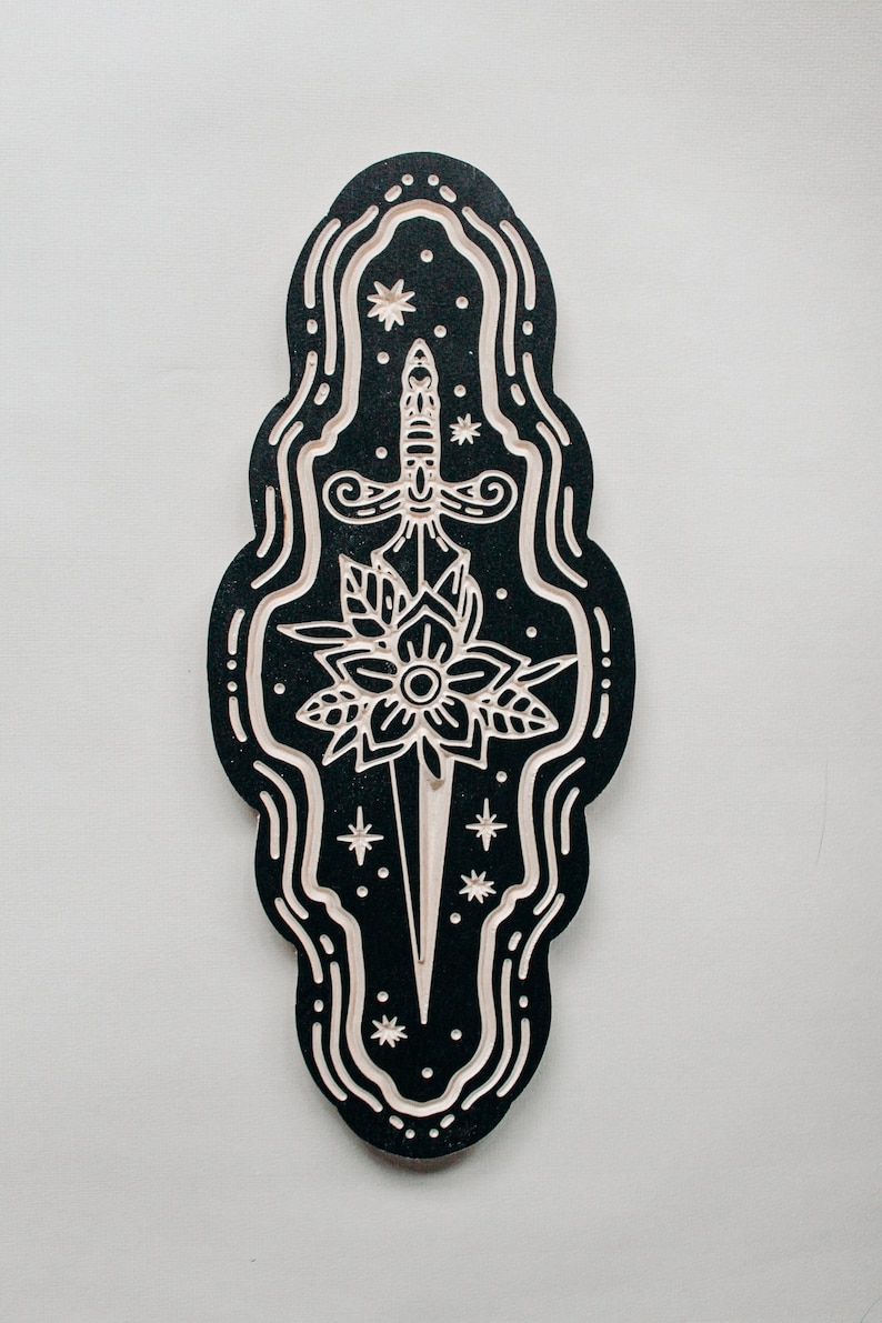 May include: A black and white wooden wall hanging with a stylized dagger and flower design. The dagger is surrounded by a swirling border and stars.