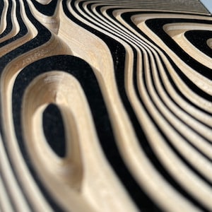 May include: Close-up of a wooden surface with a wavy, abstract pattern in black and beige. The surface is carved with a series of concentric circles and lines, creating a 3D effect.