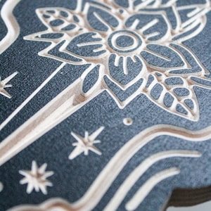 May include: A black and white wood carving of a stylized flower with a circle in the center. The flower is surrounded by a swirling pattern and stars.