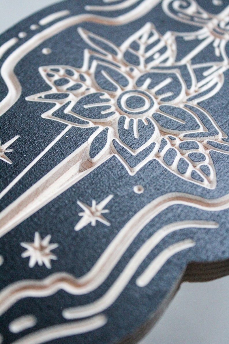 May include: A black and white wood carving of a stylized flower with a circle in the center. The flower is surrounded by a swirling pattern and stars.