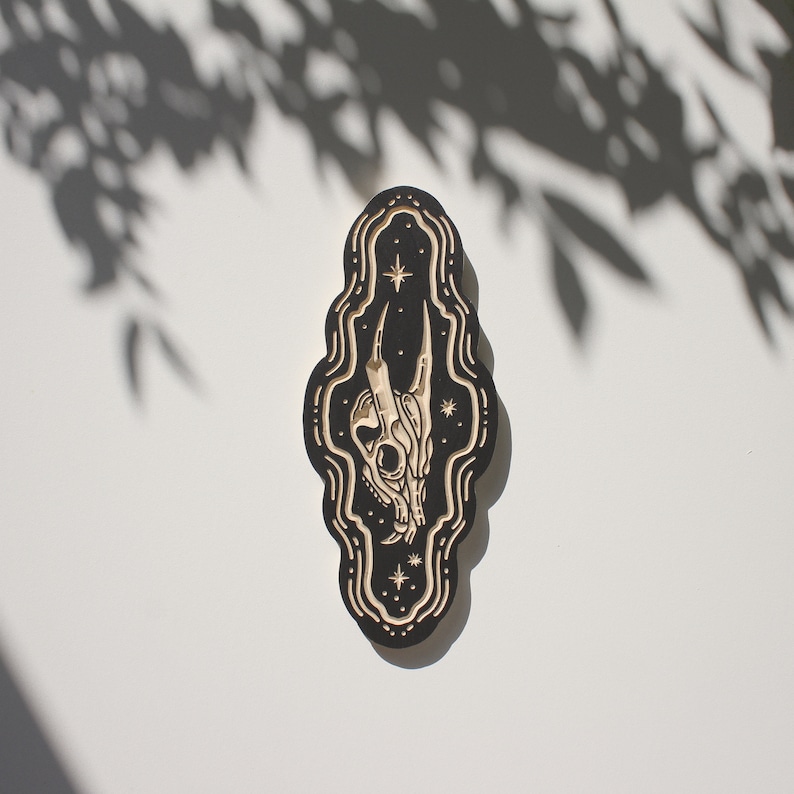 May include: A black wooden plaque with a carved design of a skull with horns and stars. The plaque is shaped like a teardrop and has a distressed look.