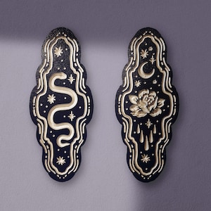 May include: Two black wooden wall hangings with gold accents. The left hanging features a snake with stars, and the right hanging features a rose with a crescent moon and dripping water.
