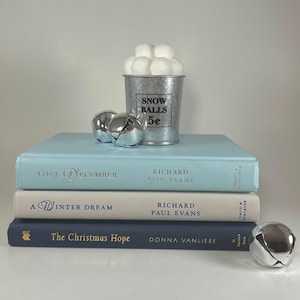 May include: A stack of three blue, beige, and dark blue hardcover books with the titles "Lost December", "A Winter Dream", and "The Christmas Hope". A small silver bucket with the words "Snow Balls 5¢" is on top of the books, filled with white felt balls. There are also three silver jingle bells on the table.
