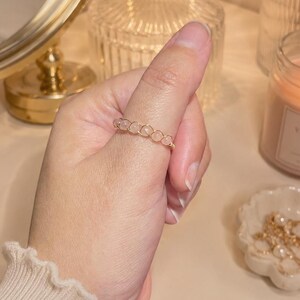 May include: A delicate ring featuring a series of small, pale pink, round beads linked together with thin, metallic gold-toned wire. The band has a woven, open-work appearance, creating a continuous pattern of circular shapes around the finger.
