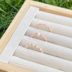 May include: Three beaded rings displayed in a wooden jewelry box. The rings feature pink, white, and gold beads. One ring has a single pearl. The rings are arranged on a light-colored fabric.