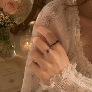 May include: Two gold rings worn on the fingers. One ring features a single round blue stone in a bezel setting. The second ring is a band composed of multiple small, round blue stones linked together in a continuous, delicate chain-like design.