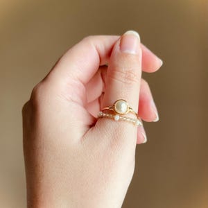 May include: Two gold-toned rings are displayed. The top ring features a large, round, off-white pearl. The bottom ring is adorned with small, clear beads. The rings are on a finger, set against a soft, neutral background.