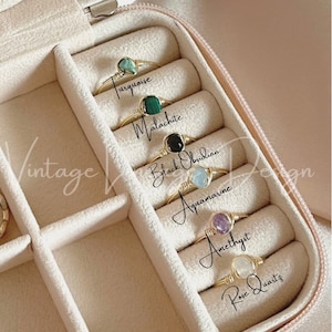 May include: A close-up of a jewelry box with six compartments, each containing a ring with a different gemstone. The gemstones are turquoise, malachite, black obsidian, aquamarine, amethyst, and rose quartz. The rings are made of gold wire wrapped around the stones.