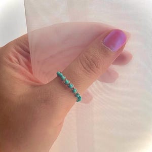 May include: A delicate ring featuring small, round, turquoise-colored beads strung together with silver-colored wire. The ring is worn on a finger, set against a soft, white background.