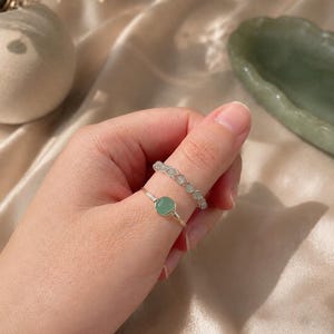 May include: Two silver-toned wire rings. One ring features a single, smooth, round, light green stone set in a wire-wrapped band. The second ring is a band composed of multiple small, light green, rounded beads linked together by silver wire.