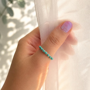 May include: A delicate ring featuring small, round turquoise beads strung on a silver-colored wire. The ring is worn on a finger, showcasing its simple yet elegant design. The turquoise beads are a vibrant contrast against the silver wire.
