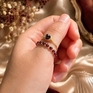 May include: Two gold-colored wire rings with red gemstones. One ring features a single, larger gemstone, while the other has a curved design with multiple smaller stones. The rings are displayed on a hand, showcasing the jewelry.
