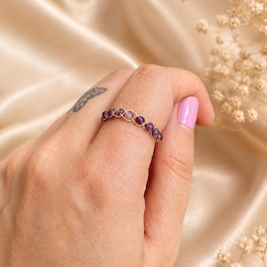 May include: A gold-colored ring featuring a series of small, round, purple gemstones. The gemstones are set in a woven pattern, creating a continuous band. The ring is displayed on a hand with a light pink nail polish.