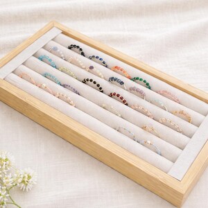 Puede incluir: Una bandeja de exhibición de joyas de madera llena de varios anillos de cuentas. Los anillos presentan una variedad de colores, incluyendo azul, rosa, verde y dorado. La bandeja tiene un forro de tela de color claro, y los anillos están cuidadosamente dispuestos.