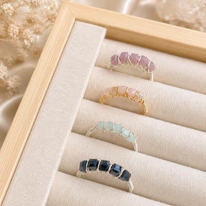 May include: A display of handmade rings with various colored gemstones. The rings are set in silver and gold wire, with gemstones in shades of pink, light blue, and black. The rings are displayed in a beige jewelry box.