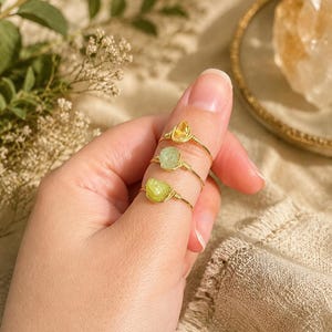 May include: Three wire-wrapped rings featuring raw, uncut gemstones in shades of yellow and green. Each ring is constructed from a thin gold-toned metal wire that secures a single, irregularly shaped stone at the center of the band.