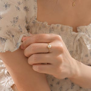 May include: A gold ring featuring a single round white pearl set in the center of a thin, polished gold band. The ring is worn on a finger, showcasing the smooth, lustrous surface of the pearl and the metallic sheen of the gold setting.