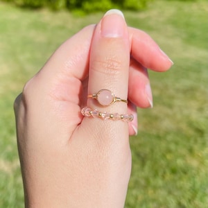 May include: Two rings on a finger. The top ring has a gold band with a round, light pink stone. The bottom ring is made of small, light pink beads and gold-colored beads. The background is blurred green.
