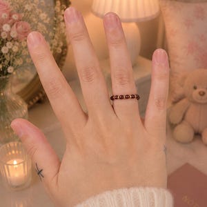 May include: A delicate ring featuring a series of small, round, deep red garnet beads alternating with tiny, polished gold-tone metal spacers. The ring is worn on the ring finger, showcasing a minimalist design with a thin, flexible band construction.