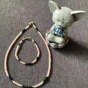 May include: A necklace and bracelet set featuring pink and black beads. The necklace and bracelet are displayed next to a gray figurine of a seated animal. The jewelry has a silver clasp.