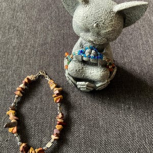 May include: A stone bracelet with a variety of earth-toned chip beads and silver-colored metal accents. A small gray statue of a creature with large ears is in the background, wearing a beaded necklace. The bracelet is approximately 7 inches long.