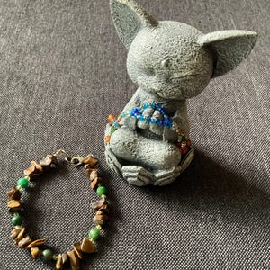 May include: A gray stone statue of a seated creature with large ears, adorned with beaded bracelets in shades of blue, orange, and green. A separate bracelet with brown and green stones is in the foreground. The jewelry is displayed on a gray surface.