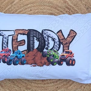 May include: White pillowcase with the name "TEDDY" printed in black, brown, and gray letters. The letters are decorated with a tire tread pattern. Four toy monster trucks are printed on the pillowcase, including a red, blue, green, and orange truck.