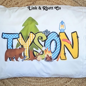 May include: A white pillowcase with the name "TYSON" spelled out in colorful letters. The letters are decorated with camping-themed designs, including a pine tree, a bear, a campfire, and a lantern. The text "Link & Rhett Co" is printed in the top right corner.