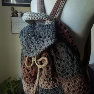 May include: A handmade crochet backpack in shades of gray, brown, and beige. The backpack features adjustable straps and a drawstring closure. The crochet pattern creates a textured appearance. The bag is displayed on a mannequin.