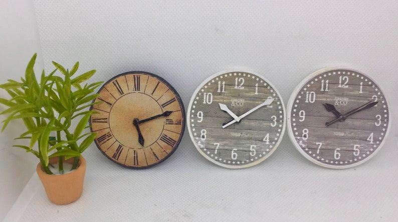 May include: Three round wall clocks with different designs. The clock on the left has a black frame and Roman numerals. The middle and right clocks have a white frame with a wood grain design and black hands. The middle clock has the text "TIMELESS & CO" on the face.