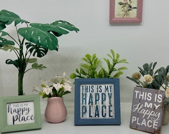 May include: Four miniature framed signs with the text "This is my happy place" in different colors and fonts. The signs are surrounded by miniature plants and a vase of white flowers.