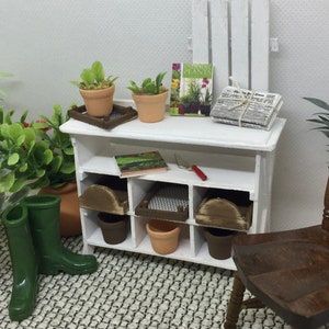 Puede incluir: Estantería de madera blanca en miniatura con un diseño de valla de piquetes en la parte superior. La estantería tiene tres estantes y está llena de artículos de jardinería en miniatura, incluyendo plantas en macetas, un libro, una pala y periódicos. La estantería está sobre una alfombra de color blanco y gris con estampado.