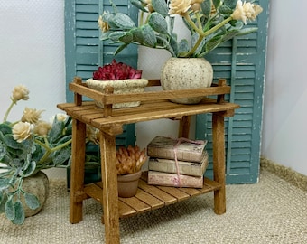 May include: A wooden two-tiered plant stand with a brown finish. The top shelf has a potted succulent plant with red flowers. The bottom shelf has a potted succulent plant and a stack of books. The stand is in front of a blue shuttered window.