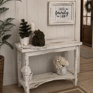 Pode incluir: Uma consola de madeira branca desgastada com uma prateleira inferior, contendo um vaso branco com flores. Uma placa emoldurada acima diz "Faith Family Farm". Os itens decorativos incluem uma pequena árvore em vaso e uma vela.