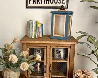 Puede incluir: Un armario de madera de estilo farmhouse con puertas de malla metálica, un letrero que dice "FARMHOUSE", una linterna azul, una pila de libros, un jarrón con flores blancas y una regadera de cobre.