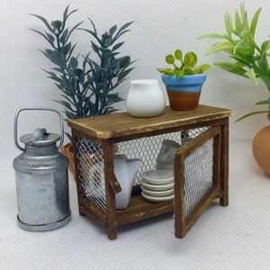 May include: A miniature wooden cabinet with a wire mesh door and shelves. The cabinet is decorated with a brown stain and has a white ceramic pitcher, a blue and brown ceramic pot with a small green plant, and a metal milk can on top.