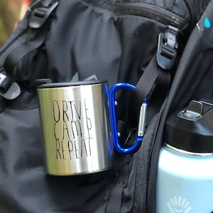 Può includere: Una tazza da viaggio in acciaio inossidabile con manico nero e coperchio nero. La tazza ha la scritta "DRIVE CAMP REPEAT" stampata sul lato. La tazza è attaccata a uno zaino nero con un moschettone blu.