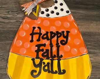 May include: A wooden candy corn door hanger with a black and white polka dot top, orange middle, and yellow bottom. The door hanger has the text "Happy Fall Y'all" written in black.