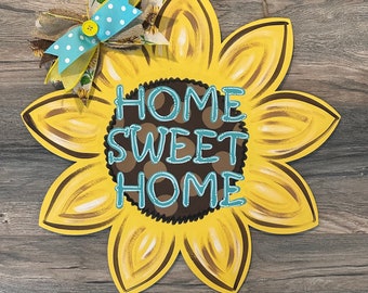 May include: A yellow sunflower door hanger with a brown center and the words "Home Sweet Home" written in blue and white letters. The sunflower has a blue and white polka dot bow on top.