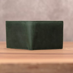 Puede incluir: Una cartera de cuero verde oscuro con un dise&ntilde;o plegado. La cartera est&aacute; sobre una superficie de madera.